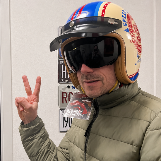 Rider flashing a peace sign in a cream open-face helmet with red and blue speed stripes, a snap-on peak visor and a dark shield
