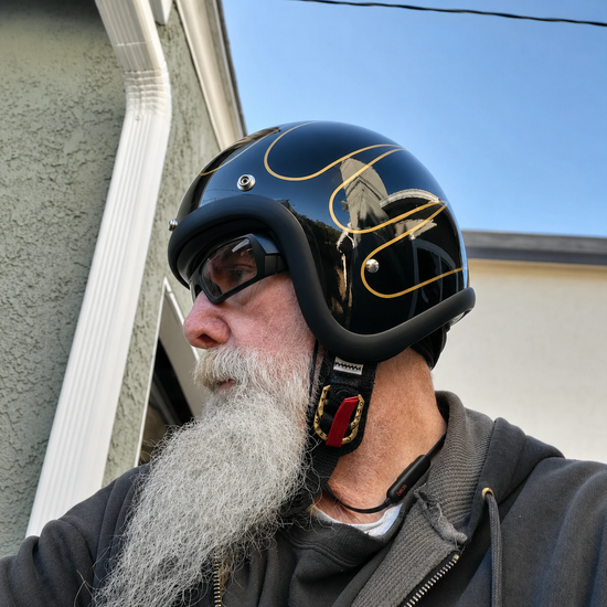 Rider with a long white beard wearing a gloss black open-face helmet with gold flame pinstripes and tinted goggles